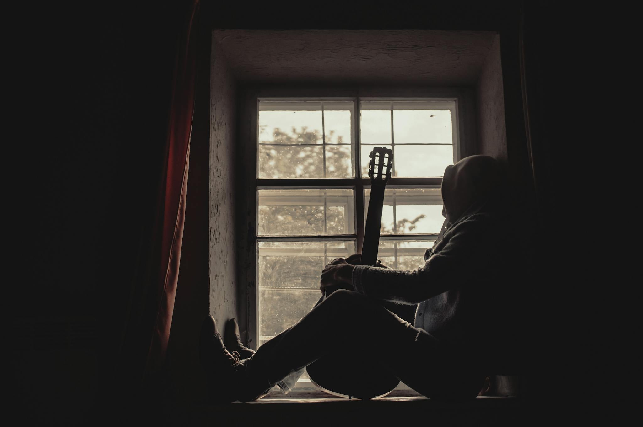 A person sits in silhouette on a windowsill with a guitar, creating a moody atmosphere.
