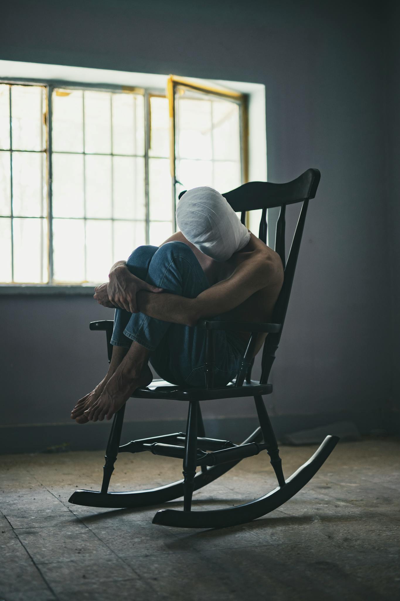 A person wrapped in cloth sits in a dark room on a wooden rocking chair by a window.