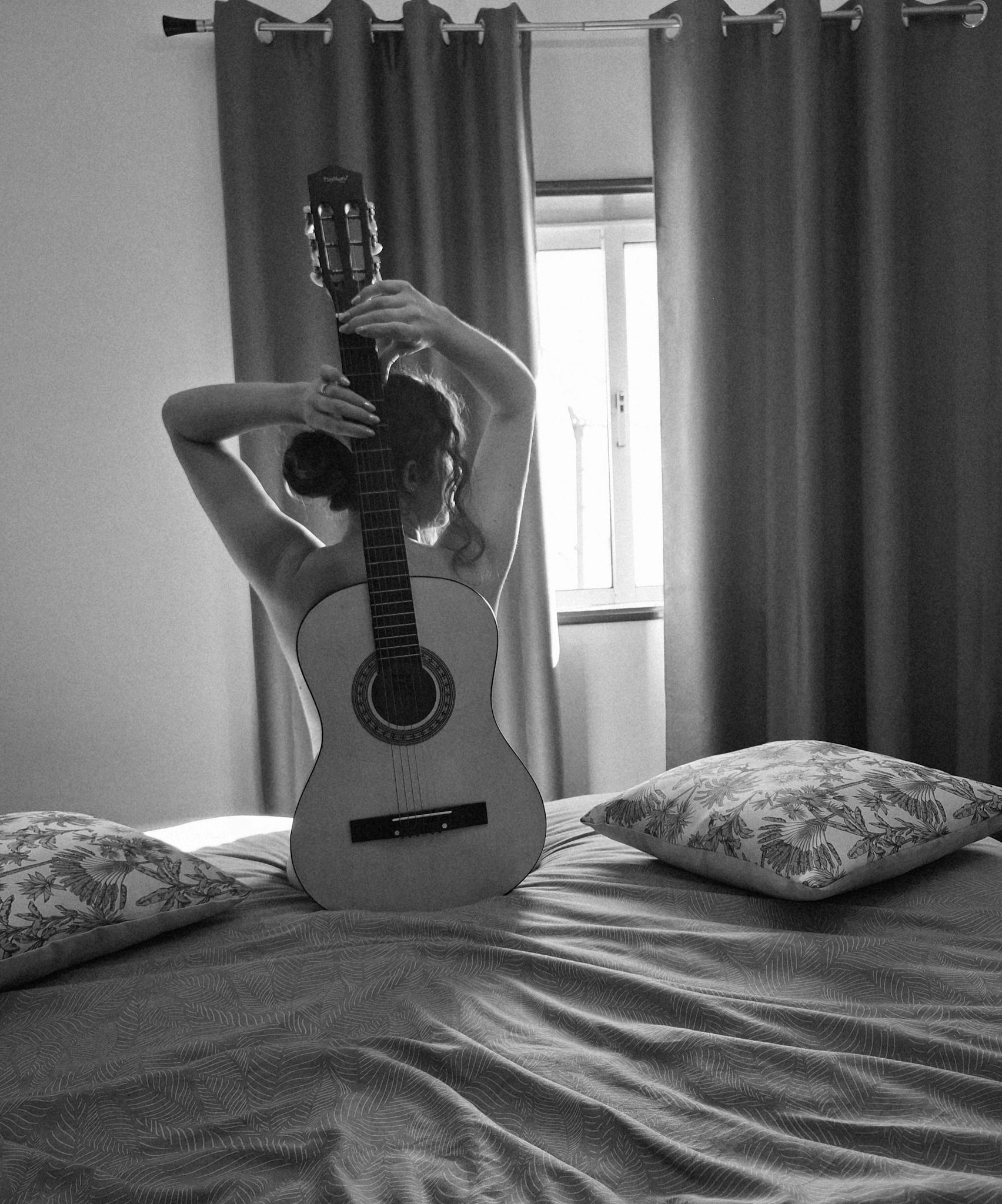 A serene black and white portrait of a person holding a guitar in a bedroom setting.