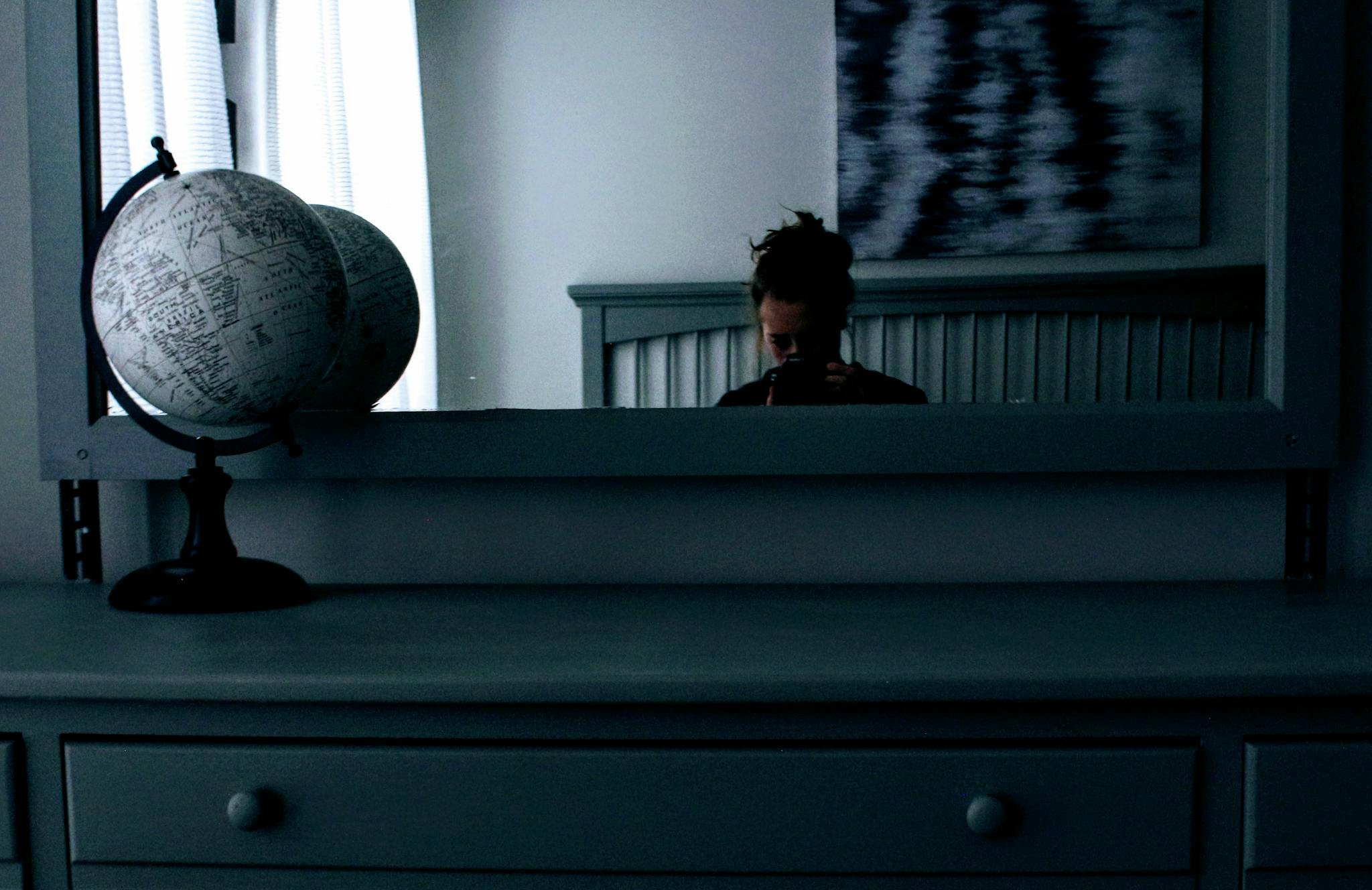A woman's reflection appears in a mirror on a dresser, with a globe beside it. Indoor setting.