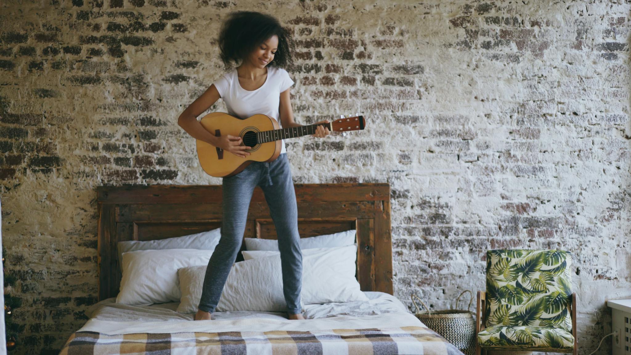 Cheerful young woman playing guitar on bed against vintage brick wall.