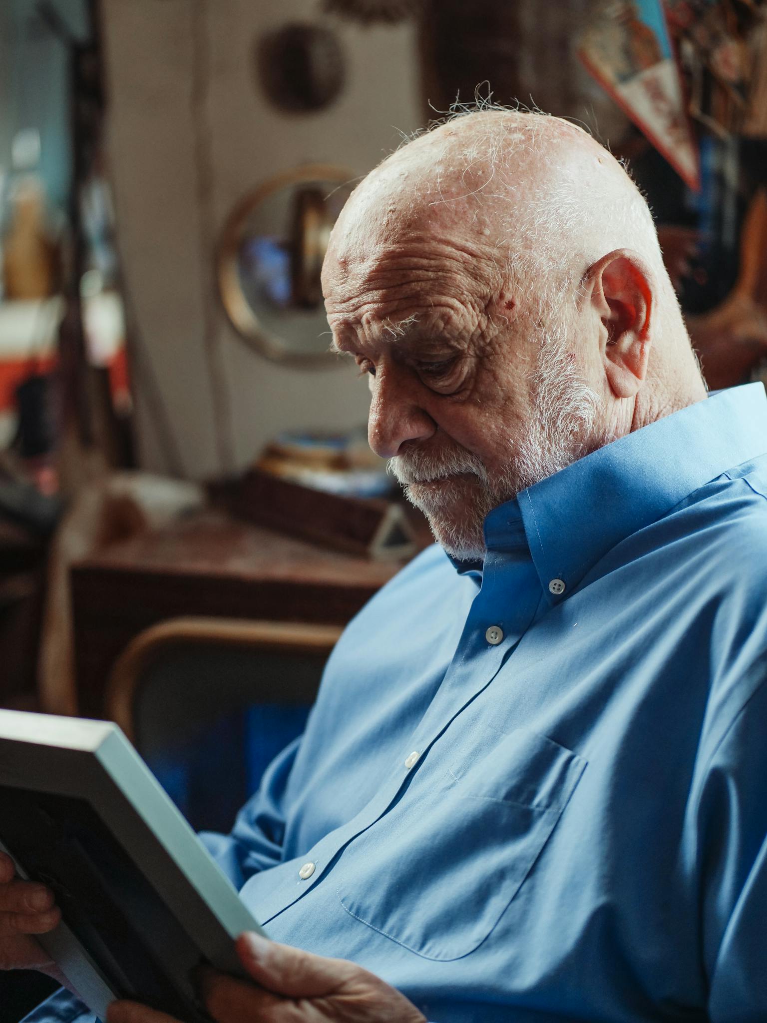 Senior man in a blue shirt sitting indoors, holding a picture frame, experiencing deep emotions.